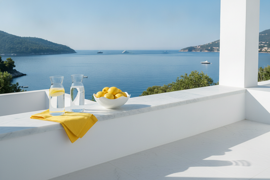 White terrace ledge with two water carafes, a bowl of lemons, and a yellow napkin overlooking a calm blue sea and hills